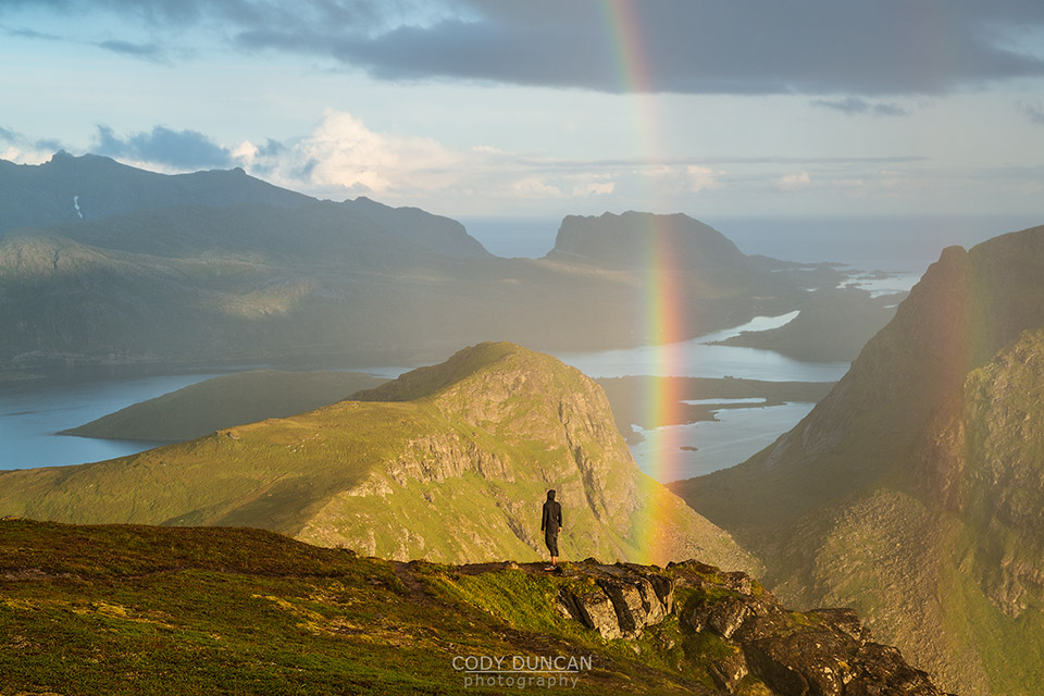 Person stands under rainbow near summit of Ryten, Lofoten Islands, Norway